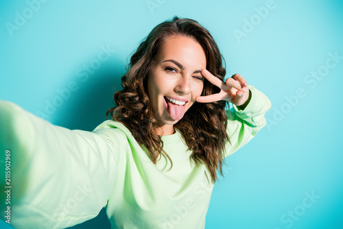 Fotografie Young woman taking a playful selfie with peace sign and tongue out against a bri