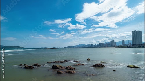 Scenic view of the rocky shoreline and modern skyline of Nha Trang, Vietnam, with calm sea, blue sky, white clouds and distant mountains along the tropical coast.