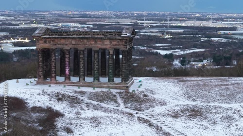 Telephoto drone orbit shot of the snow-covered Penshaw Monument in Sunderland, with the Nissan car factory and wind turbines visible in the compressed background.