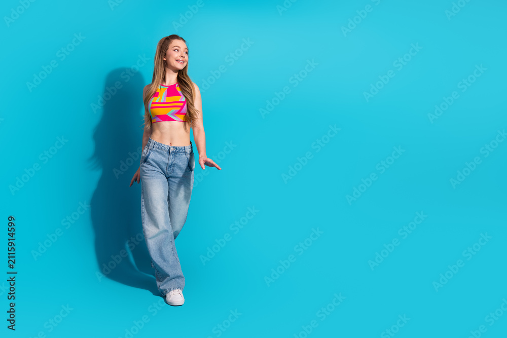 Obraz premium Young cheerful female in colorful top and jeans standing against vibrant blue background exuding happiness and style