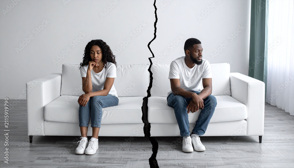 Obraz premium Sad Black couple sitting apart on a white couch split by a visual crack, symbolizing marital conflict, breakup, or divorce concept in a bright, modern living room setting.