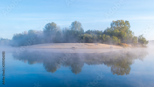 A misty morning landscape shows a serene island with lush green trees reflecting beautifully on the calm, blue river water