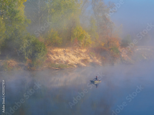 fisherman in a small boat drifts calmly on a misty river, surrounded by dense fog and colorful autumn trees lining the sandy shore