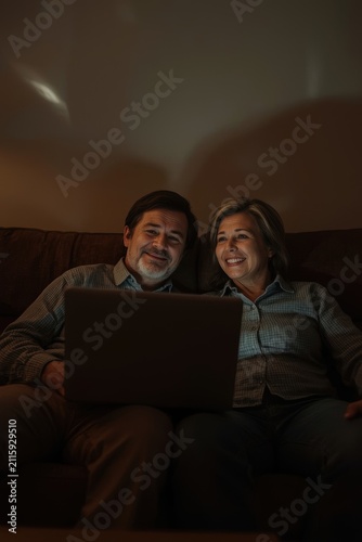 An elderly couple - a man and a woman wearing glasses - are sitting comfortably on a sofa in a semi-dark room and looking at the laptop screen with a smile.