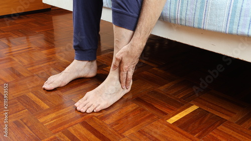 Cuadro en lienzo Elderly man painfully massages his ankle while sitting on bed, dressed in pajama pants