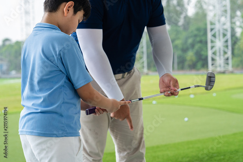 Golf Coach guiding an Asian boy through club position and grip technique during a hands-on junior golf lesson at a bright covered driving range