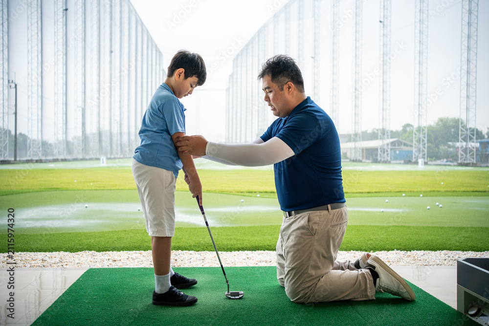 Fototapeta premium Junior Golf Lesson with a male golf coach kneeling to guide an Asian boy's arm position and stance while practicing with a club at a driving range