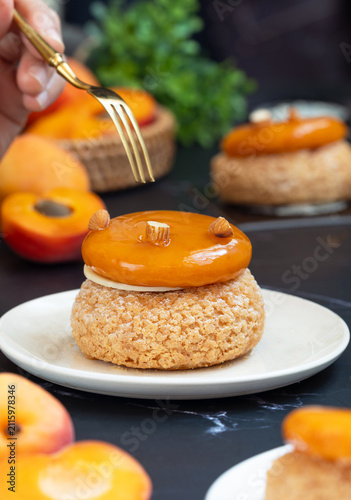 Hand holding a golden fork ready to eat French apricot peach choux au craquelin pastry served on a white plate, with apricot fruit in scene. It's a traditional pastry made with savory choux dough.