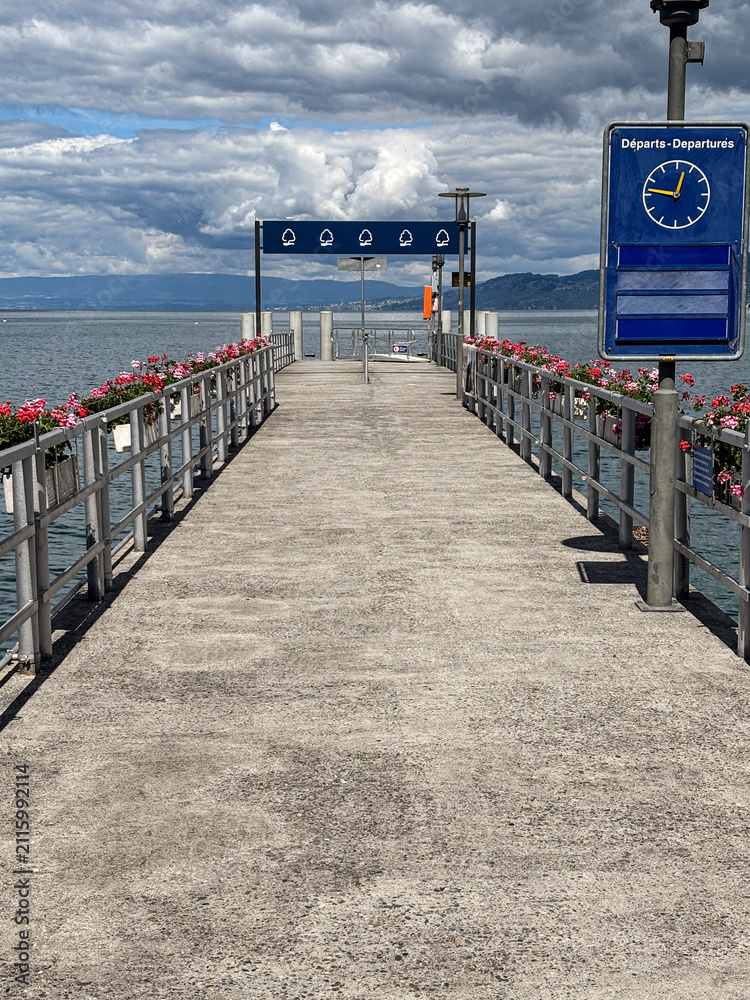 Fototapeta premium Lake geneva pier extending towards mountains under cloudy sky, decorated with vibrant flowers, showing a departure sign and clock indicating ferry travel