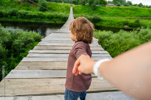 A caring mother gently guides her trusting little boy across a scenic wooden bridge over a calm lake, ensuring his safe exploration of nature while he feels secure and confident.