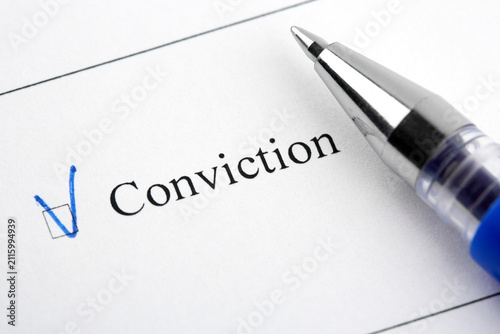 Conviction checkbox marked with a blue check mark on white paper next to a ballpoint pen. Macro shot.