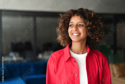 Obraz na płótnie Standing for candid portrait, curly haired latina woman using smartphone, smiling softly, glancing aside at home