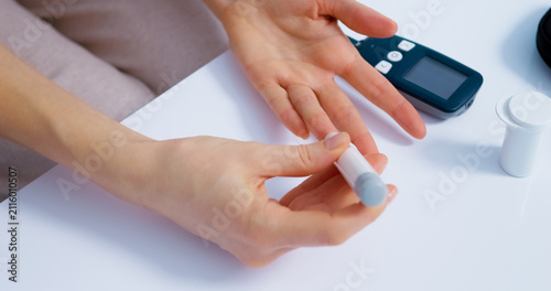 Woman prepares to check her blood sugar levels using a finger pricking device and a glucometer on a table. She focuses on her task and measures glucose.