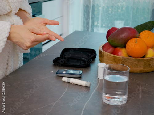 A woman performs a blood glucose test in her kitchen. She prepares to check her blood sugar level next to a bowl of fresh fruits after measuring her levels.