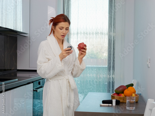 In the kitchen, a woman checks her blood sugar level with a glucometer before eating an apple. She is dressed in a robe and considers her health choices.