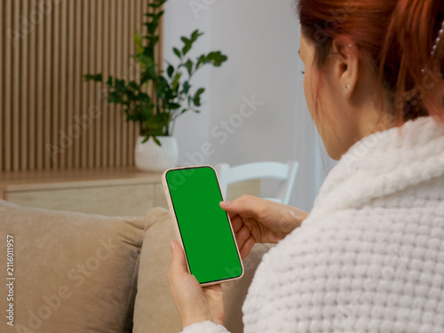 A woman sits on a sofa in the living room. She looks at a phone with a green screen. The space is cozy and has plants in the background.
