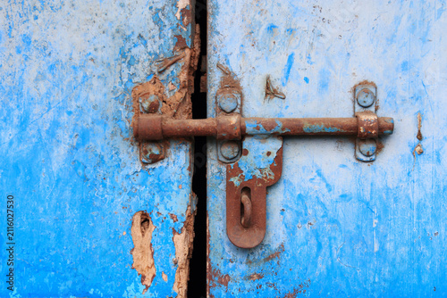 Close-up of a weathered red metal door secured with a padlock, featuring peeling paint and a rustic textured surface