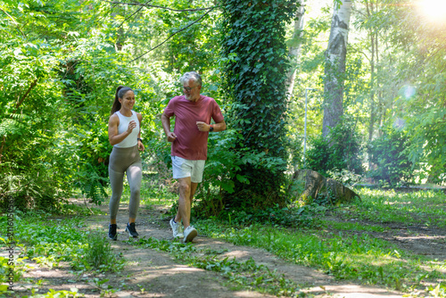 Fotografie Senior man jogging through a forest park with a fitness coach to improve cardiovascular fitness, endurance, mobility, and support healthy active aging in a natural environment