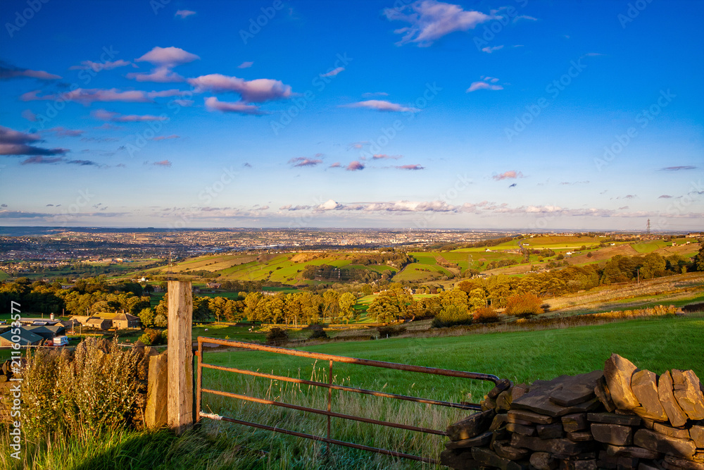 Obraz premium Panoramic golden hour landscape view over the hills and valleys of West Yorkshire, England, with the city of Bradford visible in the distance. A rusty metal farm gate and dry stone wall.