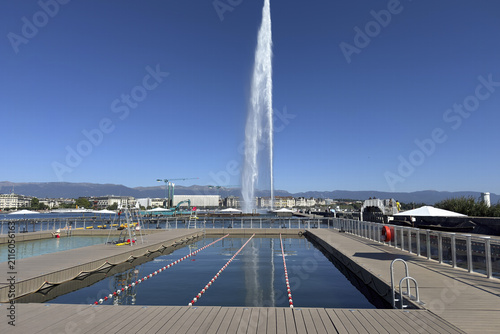 A tall jet of water rises over Lake Geneva, with people walking along the pier beneath the clear summer sky.