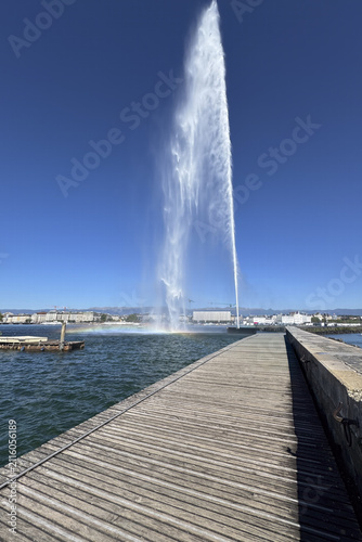 A tall jet of water rises over Lake Geneva, with people walking along the pier beneath the clear summer sky.