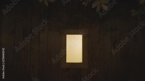 A slow camera push in towards a warm, illuminated window on the side of a rustic wooden building or cabin at night, with tree branches hanging above