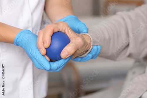 Woman therapist helping senior patient with hand rehabilitation exercise using rubber ball. Physical therapy treatment for joint pain, arthritis and bone recovery in medical facility.