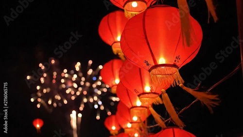 Close-up of traditional red Chinese lanterns with glowing bulbs and yellow tassels hanging outdoors at night with blurred colorful fireworks in the dark background sky during festival.