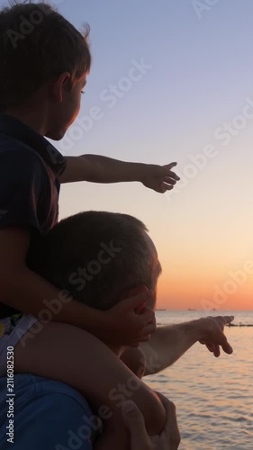 Father and son enjoy sunset at beach. Child points towards horizon while father supports him. Warm atmosphere captures bonding moment. Vertical shot highlights serene scenery.
