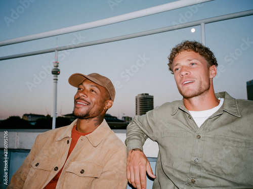 Two multiethnic male friends laughing together on a city rooftop at dusk, candid flash photo with authentic joy and urban skyline background.