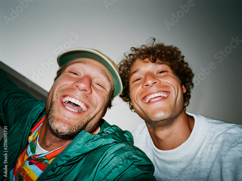 Playful wide-angle selfie of two young multiethnic male friends laughing against blue sky with construction cranes, authentic urban gen z energy.