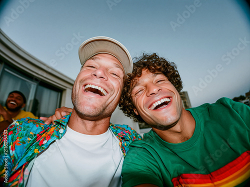 Playful wide-angle selfie of two young multiethnic male friends laughing against blue sky with construction cranes, authentic urban gen z energy.