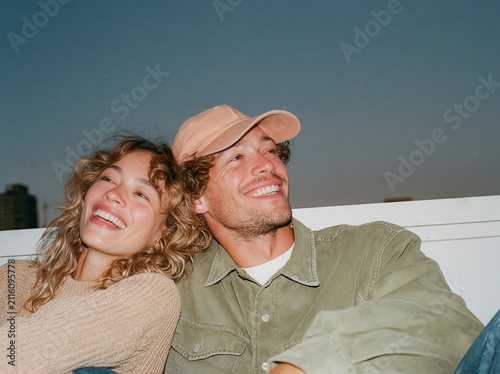 Two multiethnic male friends laughing together on a city rooftop at dusk, candid flash photo with authentic joy and urban skyline background.