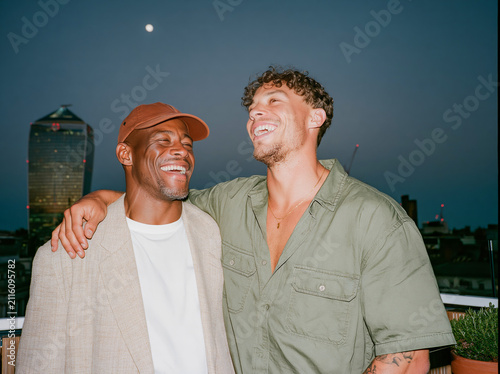 Two multiethnic male friends laughing together on a city rooftop at dusk, candid flash photo with authentic joy and urban skyline background.