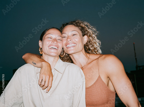 Candid flash snapshot of two happy young women embracing and laughing outdoors at night, authentic friendship moment with retro party aesthetic.