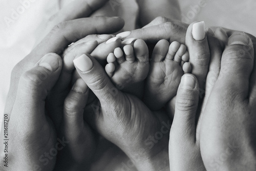 Black and white close-up of parents and older sibling holding a newborn baby's tiny feet in their hands. Family lifestyle image symbolizing love, care, bonding, protection, and togetherness.