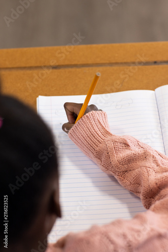 African female child writing in ruled notebook at school desk, pink sleeve holding yellow pencil