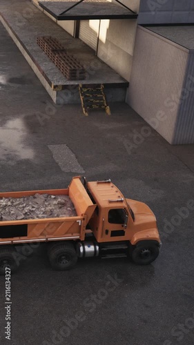 A truck is parked at a construction site. It carries building materials in its bed. Workers move bricks on the ground. The sun is shining and the area is busy with activity.
