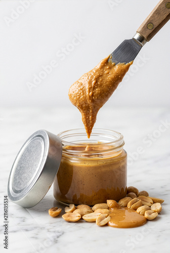 Creamy peanut butter dripping from spatula knife into small glass jar with roasted peanuts on marble surface