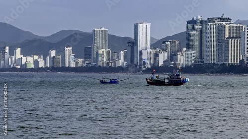 Traditional fishing boats sailing on the sea with the modern skyline of Nha Trang, Vietnam. Coastal cityscape with high-rise buildings, calm water and distant mountains.