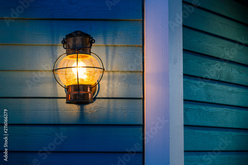 Vintage outdoor wall lantern glowing on blue wooden house exterior at dusk.