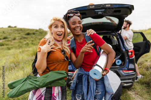 Two cheerful women stand by their car, excited for a camping trip. They wear casual outfits and hold camping gear, surrounded by grassy hills under bright sunlight.