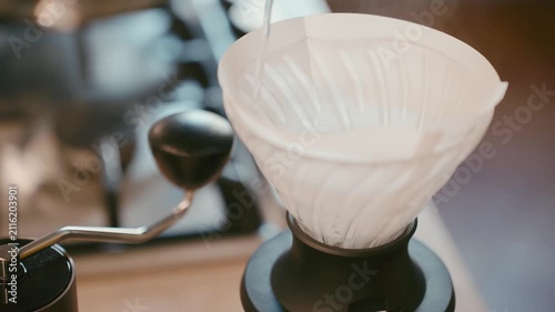 Pour over coffee brewing process, pouring hot water into ceramic dripper with paper filter, manual coffee grinder in background, specialty coffee preparation at home