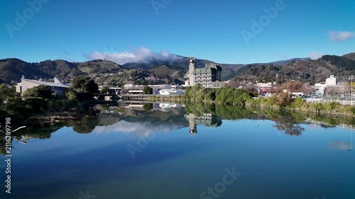 Nelson, New Zealand - 14 July 2026: Aerial view of Civic House reflected in the calm waters of the Maitai River, with surrounding hills and a clear blue sky.