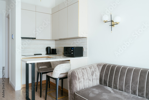 Contemporary home interior showcasing a minimalist kitchen design with white cabinetry, a bar counter, and comfortable bar stools, alongside a stylish grey velvet sofa in an open plan living space