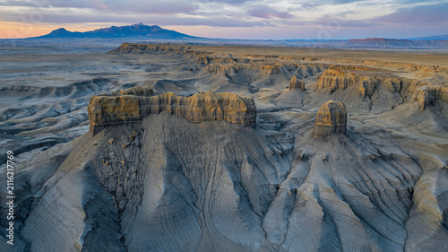 Aerial view of the rugged badlands landscape with towering sandstone formations, deep erosion gullies, and distant mountains under a soft sunset sky in Hanksville, Utah, United States.