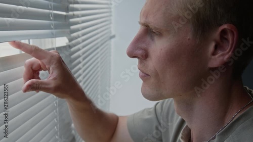 Caucasian young adult man wearing dog tags looking through window blinds with serious expression. Footage for mental health awareness campaigns, PTSD support, or security services