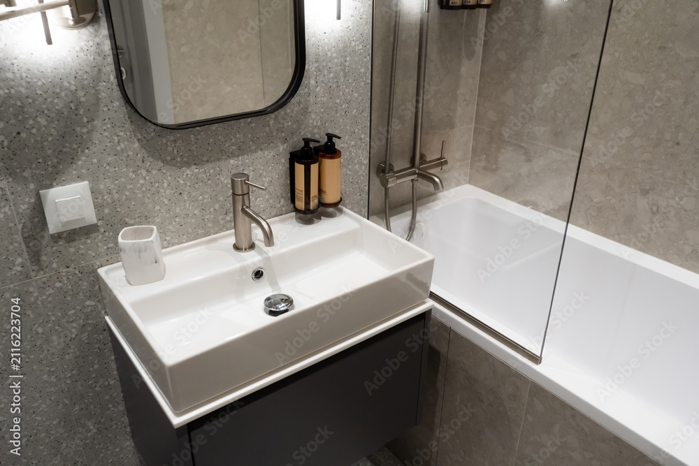 Fototapeta premium Contemporary bathroom featuring a stylish vanity sink, sleek faucet, and white bathtub, surrounded by gray terrazzo and large format tiles, reflecting modern design and comfort
