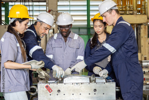 Diverse factory workers and engineers brainstorming around industrial machinery mold.Team of multiethnic industrial engineers discussing work project in factory.Group of diverse technicians examining 