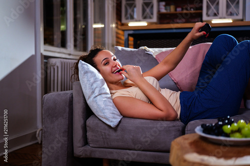 Happy young woman laying comfortably on a sofa at home during evening, enjoying leisure time by eating grapes and watching television with a remote control